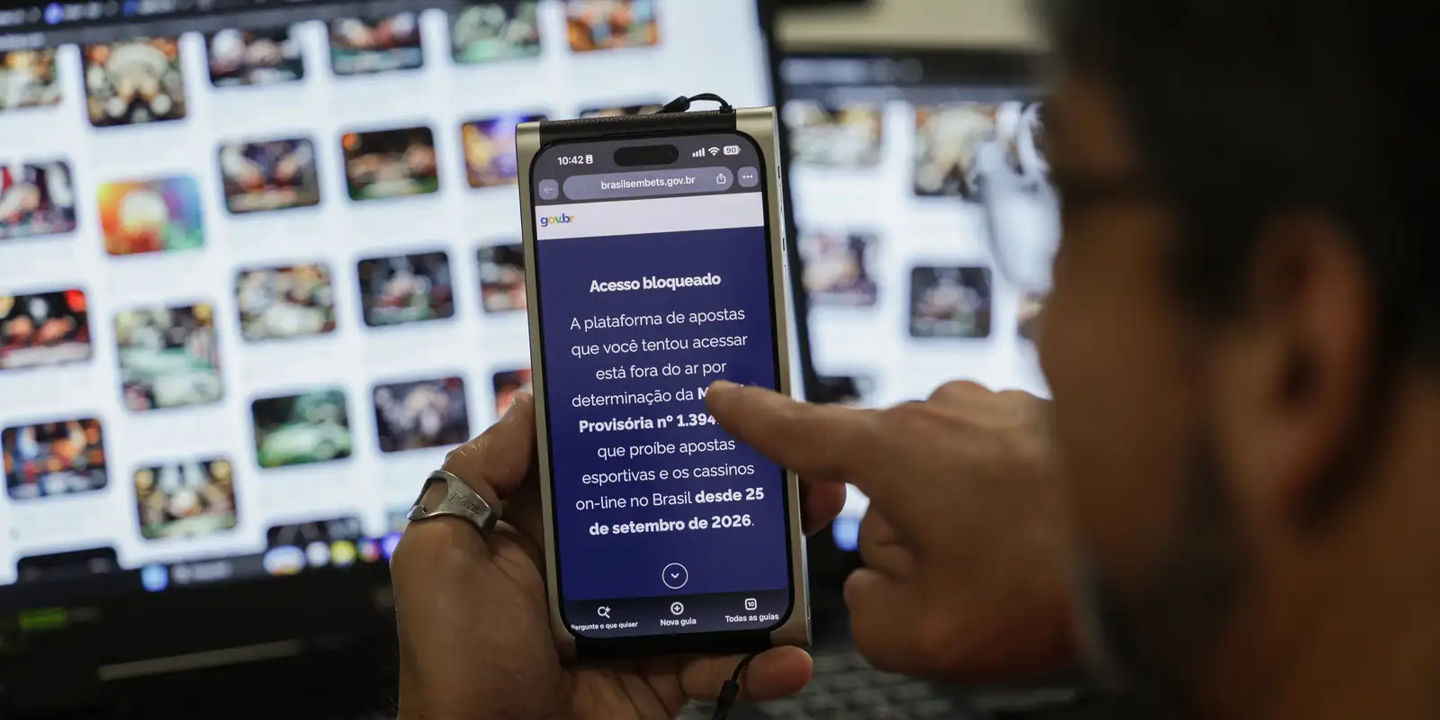Mão segurando celular com a mensagem de acesso bloqueado a plataforma de apostas, com uma tela de jogos on-line ao fundo