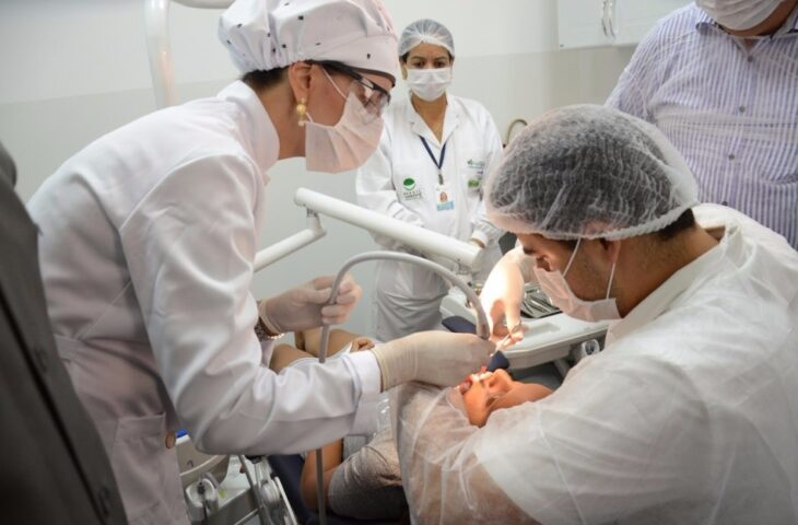 Dentistas de máscara e touca examinam a boca de uma paciente deitada em cadeira odontológica