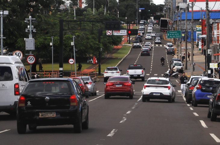 Carros em fila em avenida movimentada, com semáforos e placas de trânsito