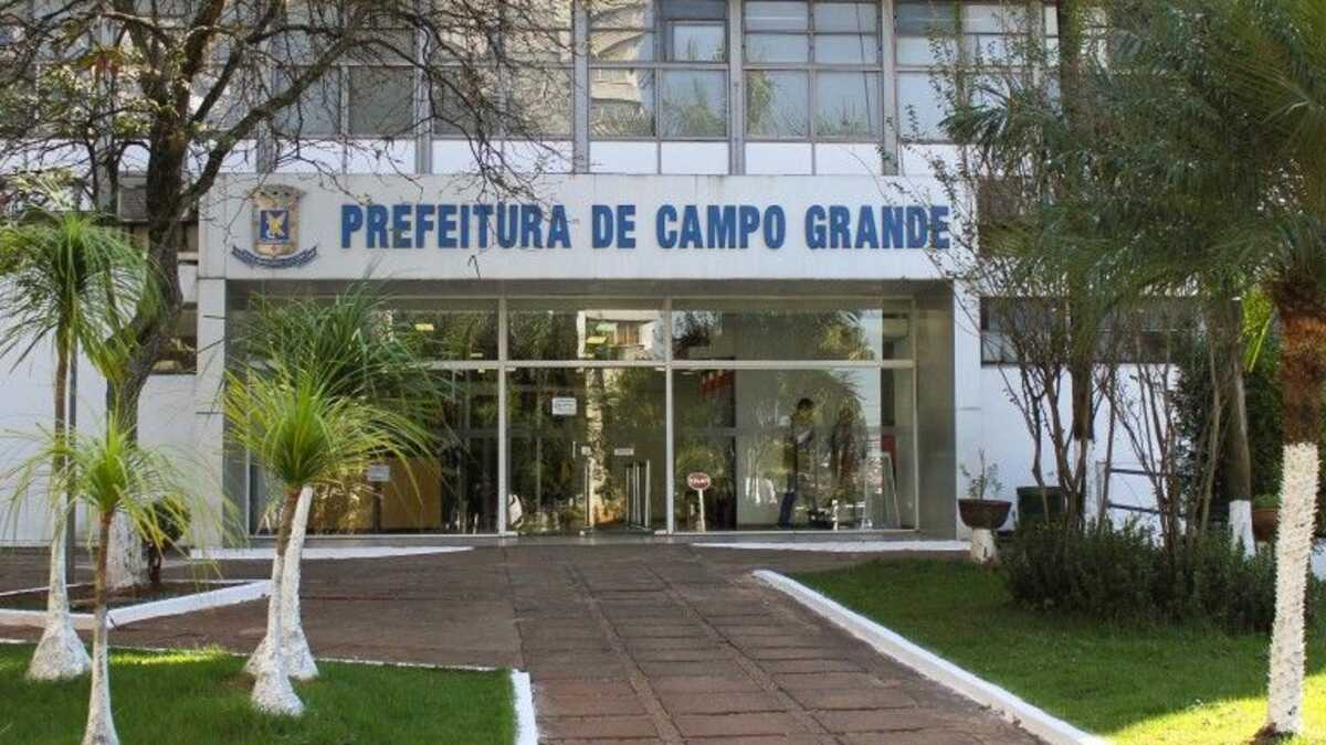 Fachada do prédio da Prefeitura de Campo Grande, com letreiro azul, entrada de vidro e palmeiras no jardim