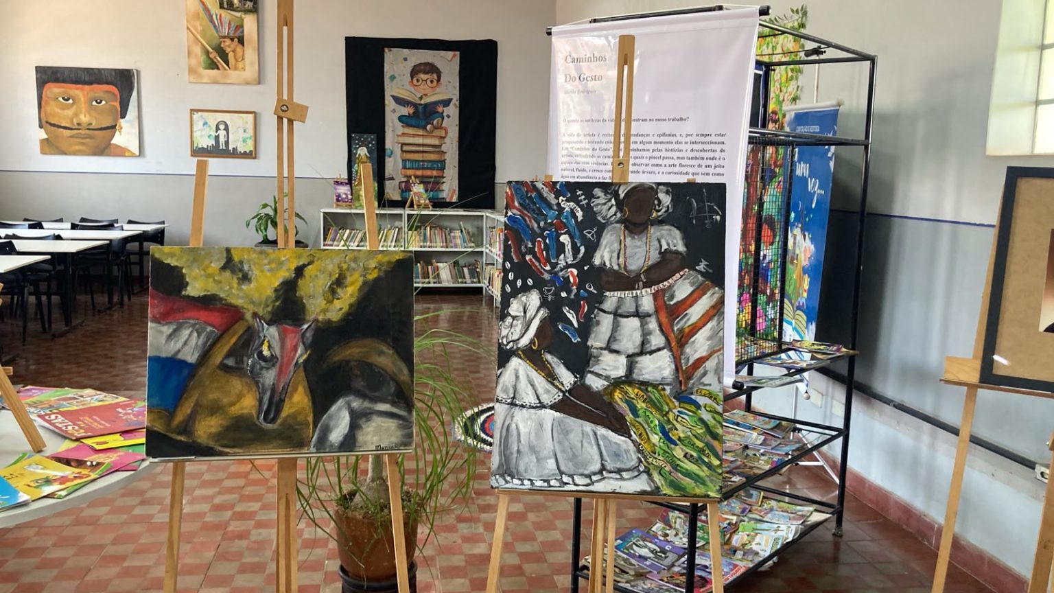 Quadros coloridos em cavaletes, entre eles duas baianas e uma figura com faixas verde e amarela, numa sala da biblioteca com estantes ao fundo