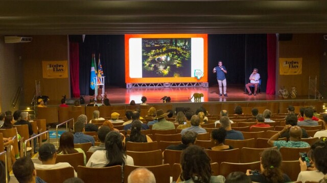 Auditório com plateia sentada assistindo a uma apresentação projetada em telão no palco
