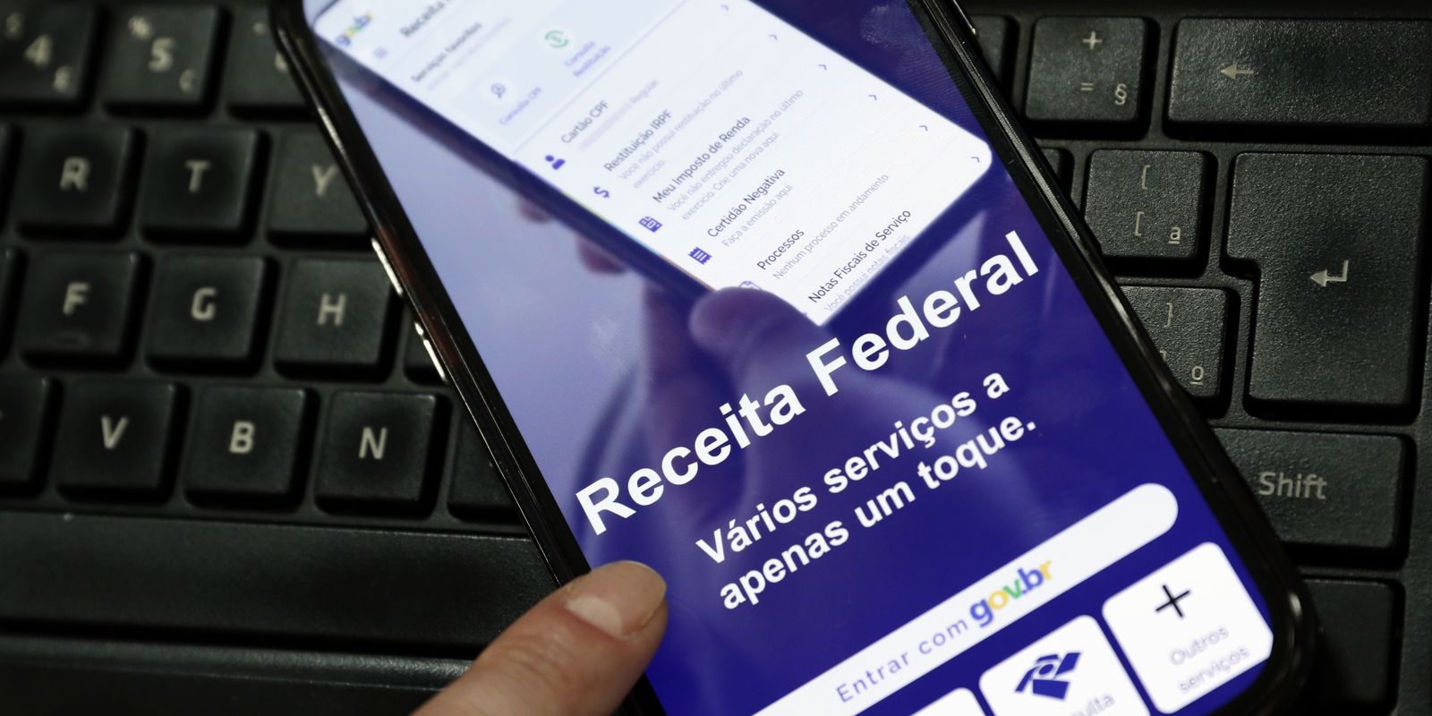 Mão segura celular com a tela do aplicativo da Receita Federal sobre um teclado de computador