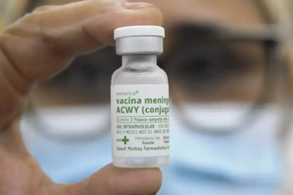Mão segura frasco da vacina meningocócica ACWY diante de profissional de saúde de máscara, fora de foco