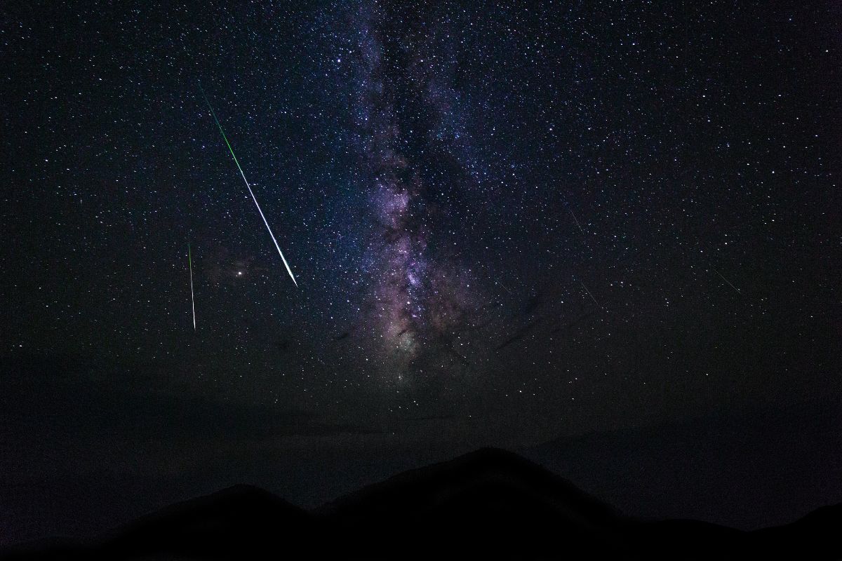Rastro luminoso de meteoro cruzando céu estrelado com a Via Láctea sobre silhueta de montanhas