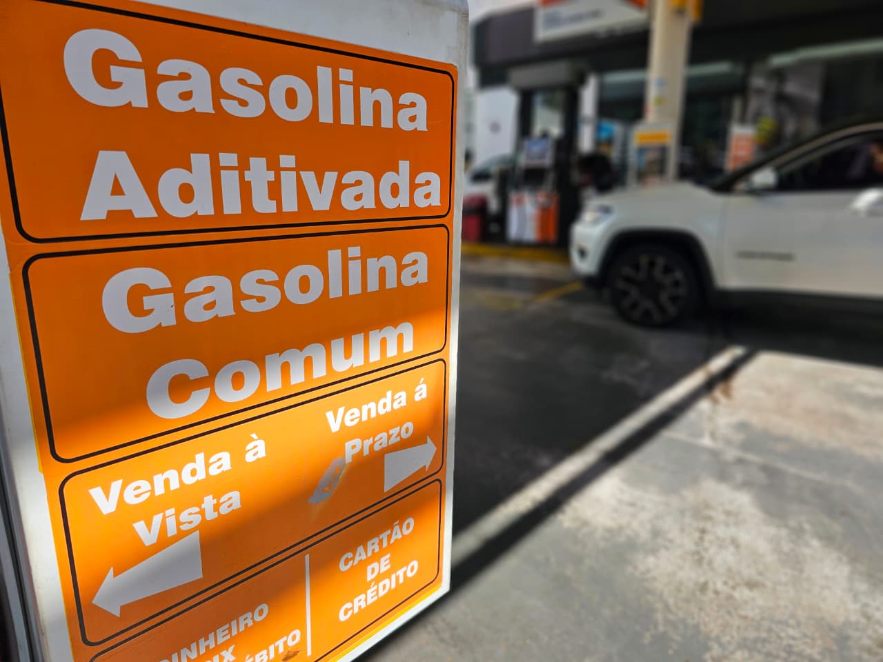 Placa laranja de posto indicando gasolina aditivada e comum, venda à vista e a prazo, com carro branco ao fundo