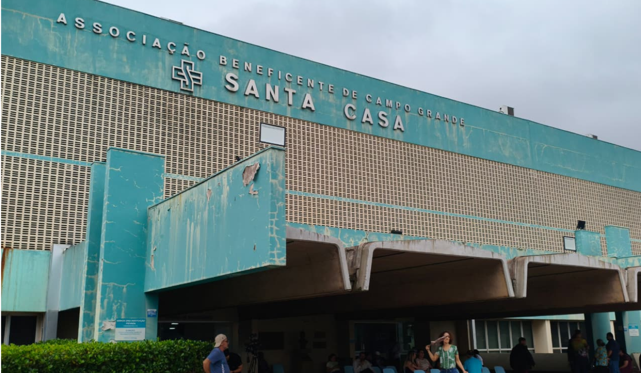 Fachada azul-esverdeada da Santa Casa de Campo Grande, com letreiro e pessoas na entrada