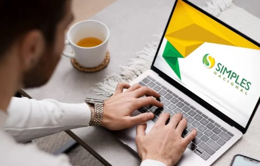 Pessoa digitando em notebook com a marca do Simples Nacional na tela, ao lado de uma xícara de chá