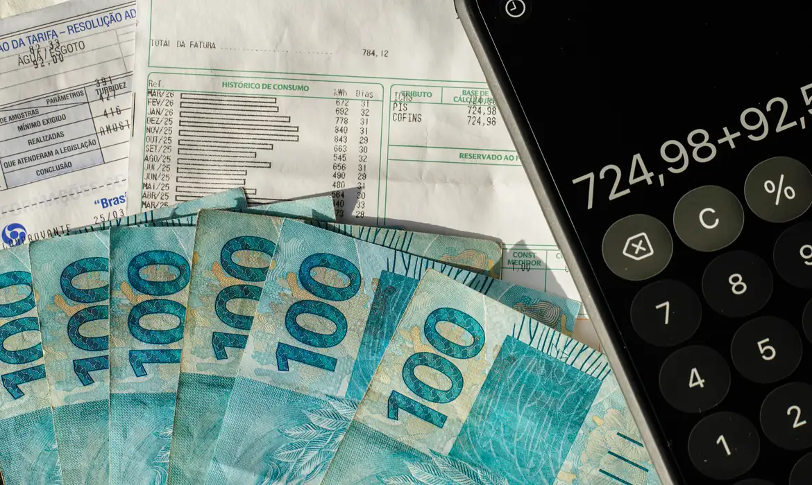 Cédulas de R$ 100 espalhadas sobre faturas com campos de tributos PIS e Cofins, ao lado de celular com calculadora aberta