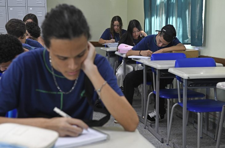 Estudantes de uniforme azul escrevendo em cadernos numa sala de aula