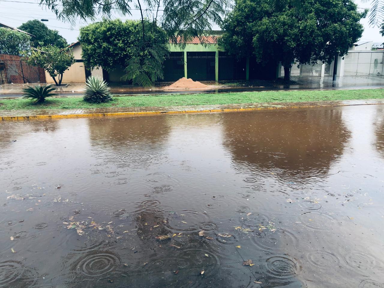 Rua alagada sob chuva em Aquidauana, com casas e árvores ao fundo