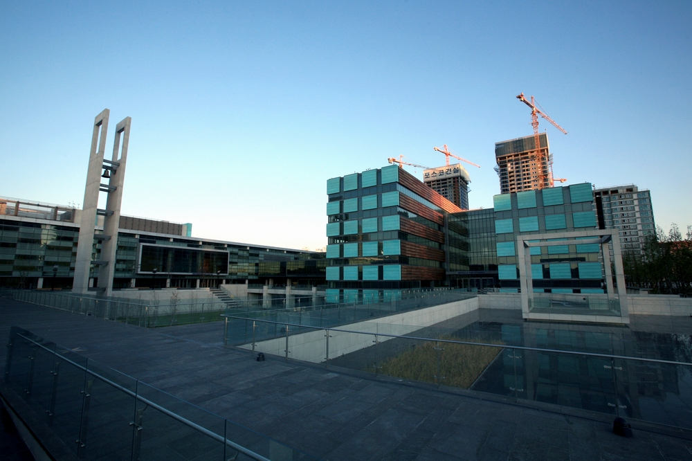 Vista externa do campus da Chadwick International em Songdo, com prédios de fachada envidraçada em painéis azul-turquesa e cobre, torre de sino em concreto e espelho d'água sob céu azul