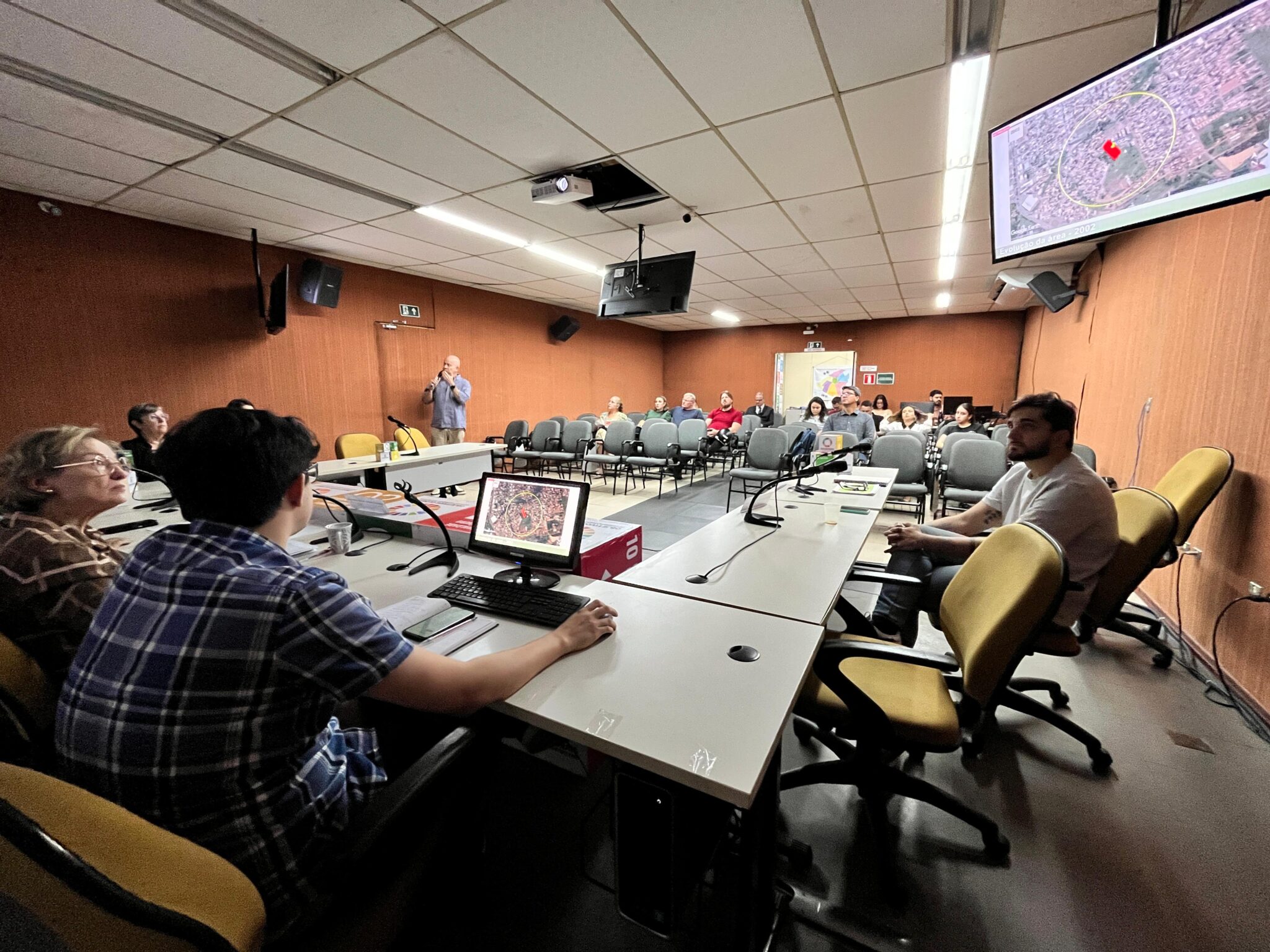 Sala de reunião com pessoas sentadas diante de mesas e telão exibindo mapa de um bairro