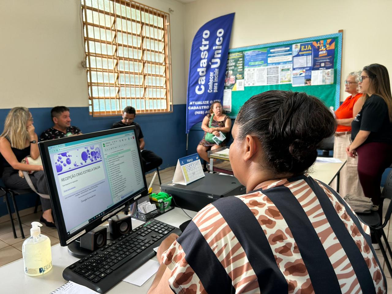 Mulher de costas diante de computador em atendimento do Cadastro Único, com banner do programa ao fundo