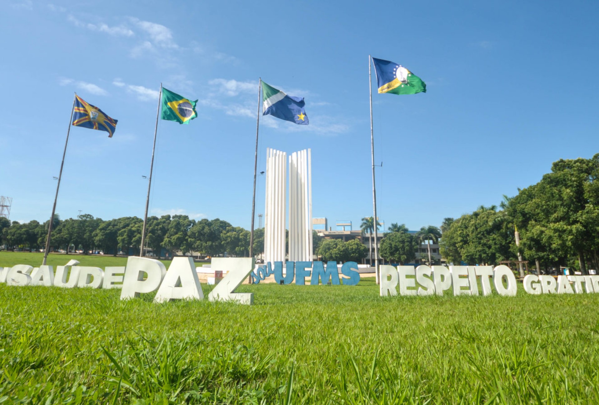 Gramado com letreiros brancos e o nome UFMS, mastros com bandeiras e o monumento branco da Cidade Universitária sob céu azul