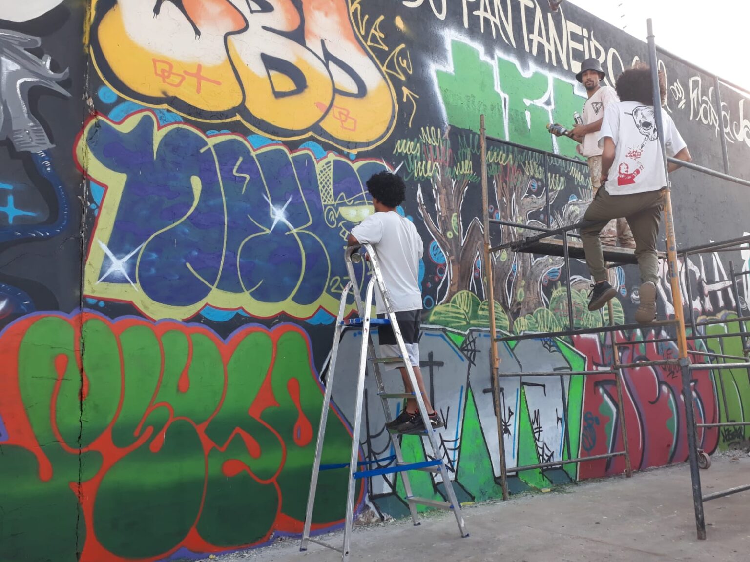 Artistas pintam grafite colorido em muro com andaime e escada, em imagem de divulgação da Prefeitura de Campo Grande usada na divulgação do edital de muralismo