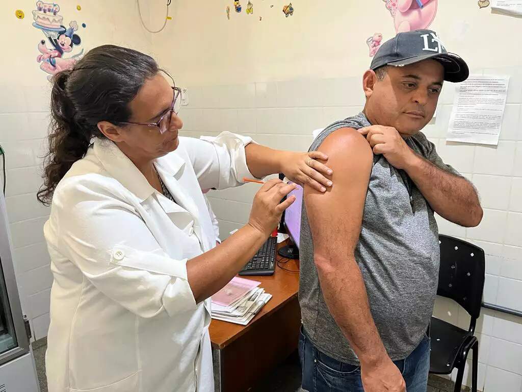 Profissional de saúde de jaleco branco aplica vacina no braço de um homem de boné em uma sala de vacinação