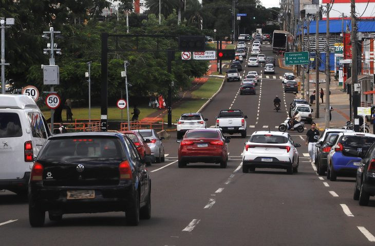 Carros em fila em avenida movimentada, com semáforos e placas de trânsito