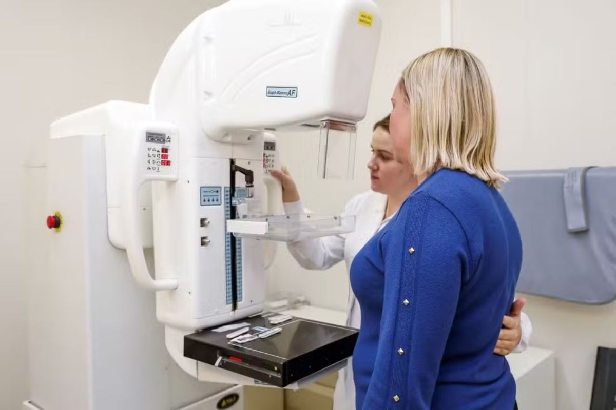Técnica em radiologia posiciona paciente diante de um mamógrafo em sala de exames