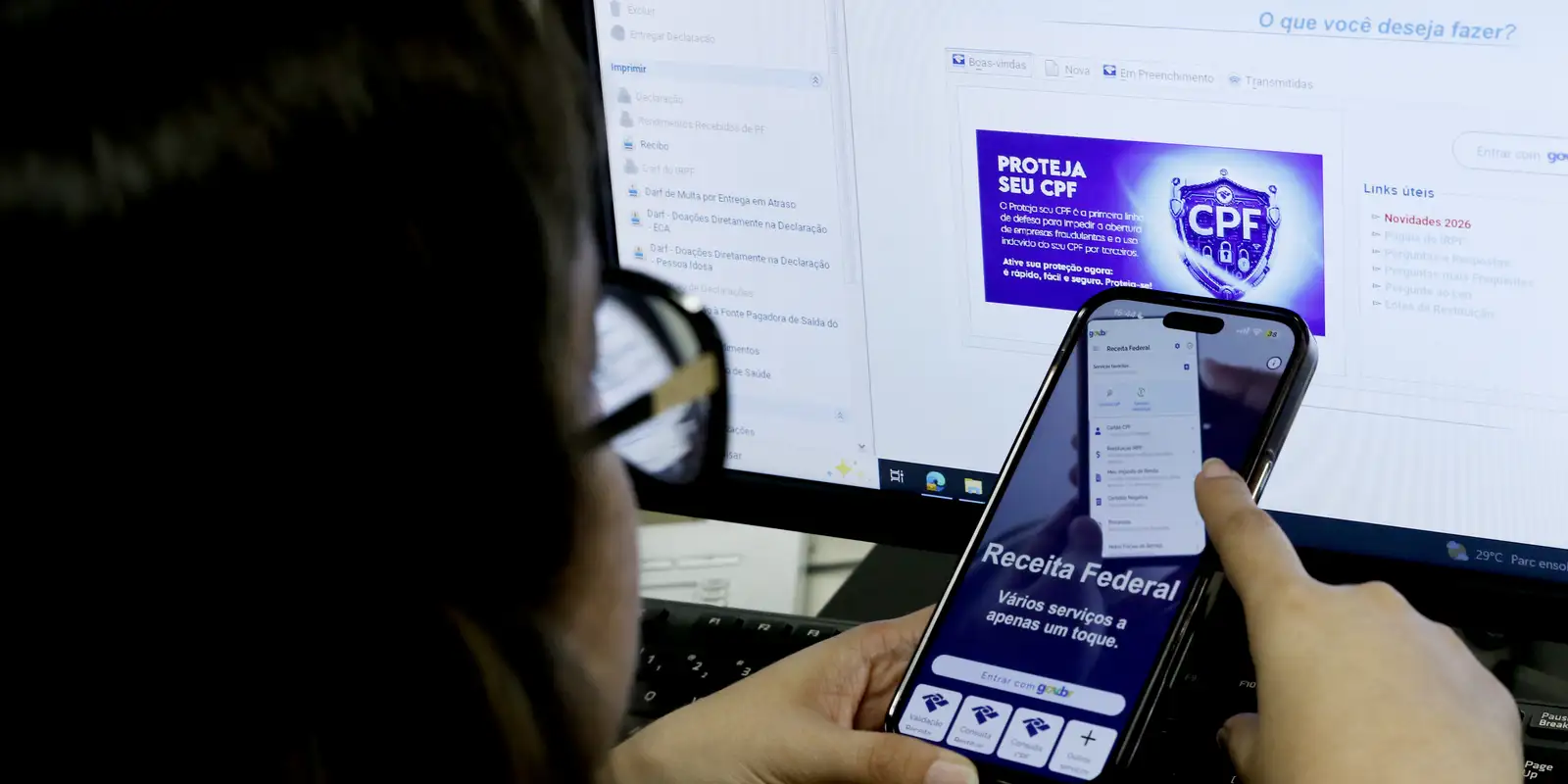 Pessoa usa o aplicativo da Receita Federal no celular diante de um computador com o site do órgão aberto