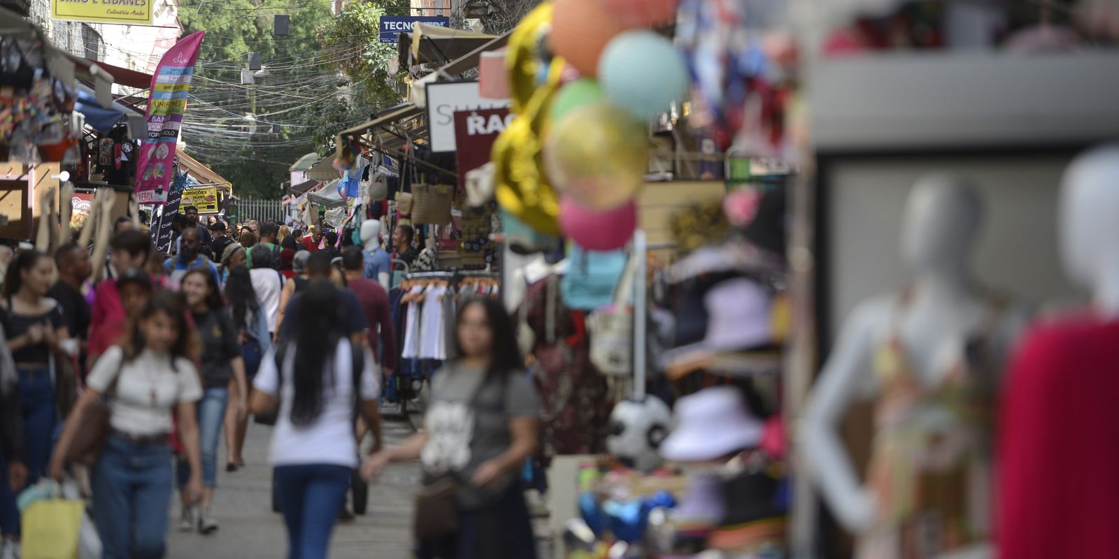 Rua estreita de comércio popular cheia de pedestres, com lojas de roupas e manequins expostos na calçada