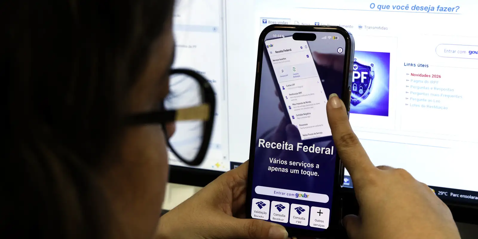 Pessoa segura celular com o aplicativo da Receita Federal aberto, diante de monitor com a página do Imposto de Renda