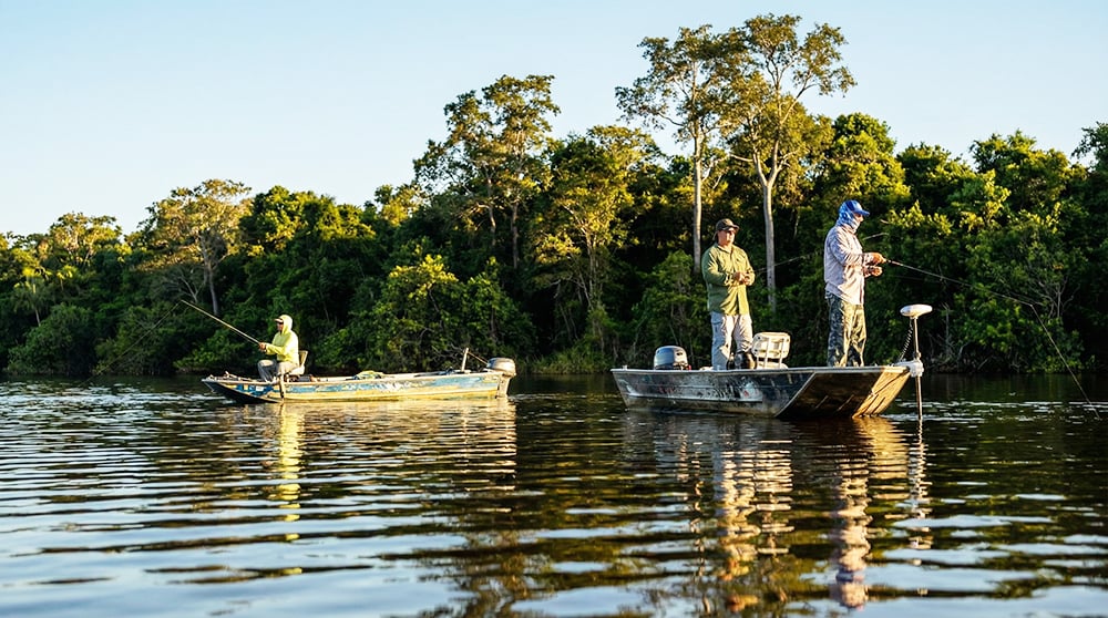 Pescadores em dois barcos de alumínio num rio, com mata ciliar ao fundo, no fim de tarde