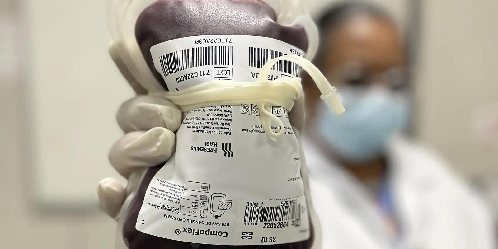 Mão com luva segurando bolsa de sangue etiquetada, com profissional de máscara desfocado ao fundo