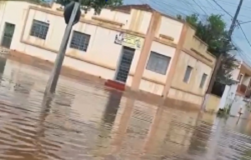 Rua alagada sob chuva em Aquidauana, com casas e árvores ao fundo