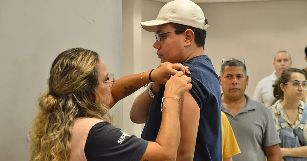 Profissional de saúde com camiseta do SUS aplica vacina no braço de um jovem de boné enquanto outras pessoas aguardam na fila